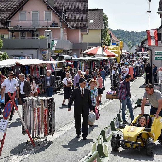Ein geschäftiger Straßenmarkt mit verschiedenen Ständen, die Waren verkaufen. Menschen gehen die Straße entlang, einige tragen Taschen. Ein Mann in einem Anzug geht mit einer Frau, und ein Kind fährt einen gelben Go-Kart. Gebäude säumen die Straße, und Berge sind in der Ferne sichtbar.