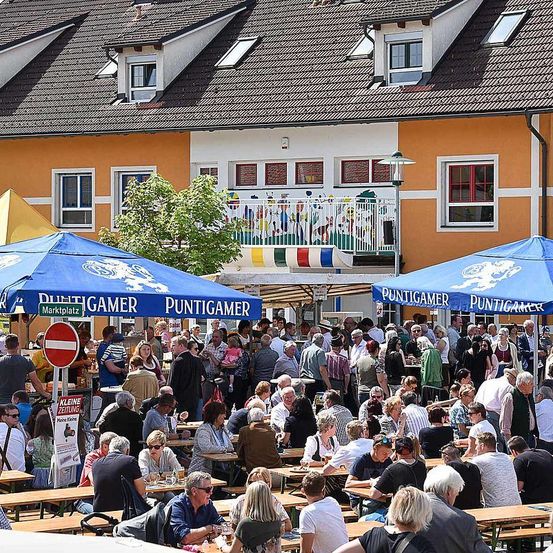 Eine Freiluft-Versammlung unter großen blauen Zelten auf einem Markt, mit vielen sitzenden und stehenden Personen, wahrscheinlich bei einem Festival. Der Bereich ist gefüllt mit Tischen und Stühlen, und das Gebäude dahinter hat einen Balkon mit farbenfrohen Dekorationen.