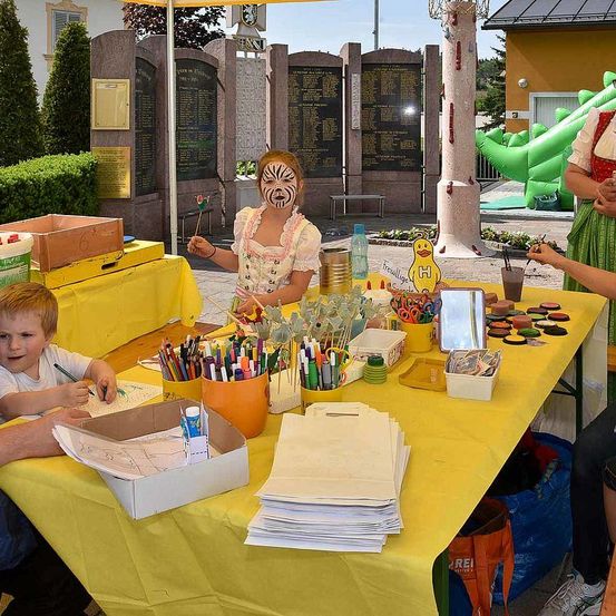 Kinder malen an einem Tisch mit gelber Tischdecke, umgeben von Malutensilien. Ein Kind hat Tiger-Gesichtsbemalung. Dahinter hilft ein Erwachsener einem Kind beim Zeichnen. Im Hintergrund steht ein Zaun mit Tafeln und ein Spielplatz.