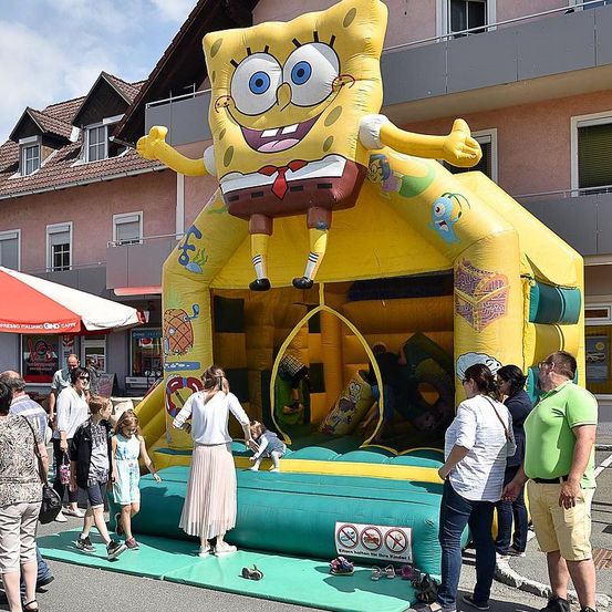 Menschen versammeln sich um einen großen aufblasbaren SpongeBob-SquarePants-Hüpfburg auf der Straße. Kinder spielen im Inneren, während Erwachsene beobachten. Hinter der Hüpfburg befindet sich ein Gebäude mit Balkon.