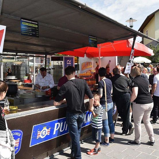 Ein Imbissstand mit einem roten Sonnenschirm hat ein Schild mit der Aufschrift "PUMPERNIKEL". Ein Mann in einem weißen Hemd kocht hinter einem Tresen. Mehrere Leute stehen in der Schlange, darunter ein Kind. Eine Frau schaut sich das Essen an.