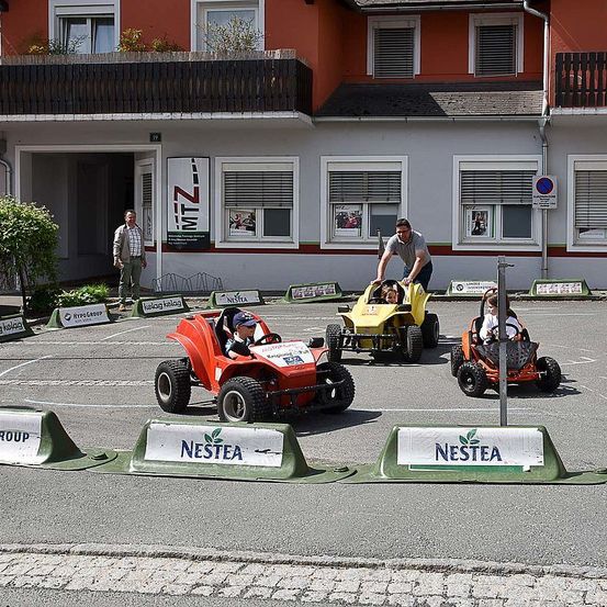 Vier Go-Karts treten auf einer Rennstrecke vor einem Gebäude an. Zwei Männer beaufsichtigen das Rennen.