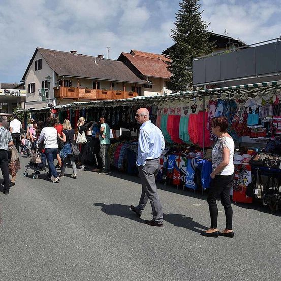 Ein geschäftiger Straßenmarkt an einem sonnigen Tag, mit verschiedenen Ständen, die Kleidung und Accessoires verkaufen. Menschen schlendern herum, einige sind einkaufen. Im Hintergrund befinden sich Gebäude mit Balkonen und eine hohe Tanne.
