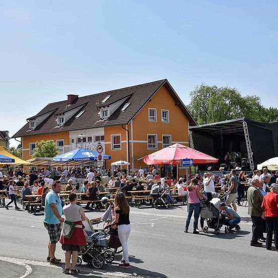Ein geschäftiger Straßenmarkt unter blauen und roten Schirmen, mit Menschen, die an Tischen sitzen und essen. Ein gelbes Haus mit einer Bühnenaufstellung ist sichtbar, und die Menge ist versammelt, um eine Aufführung zu sehen.