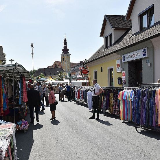 Eine Außenmarktstraße mit verschiedenen Ständen, die bunte Kleidung verkaufen, und einem Mann, der vor ihnen steht. Gebäude mit Schildern befinden sich auf beiden Seiten. Ein Turm ist in der Ferne sichtbar.