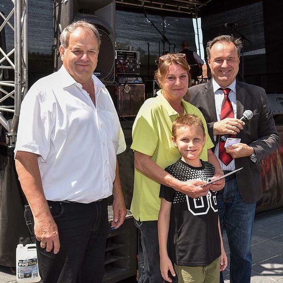 Drei Erwachsene und ein Junge stehen zusammen vor einer Bühne mit Lautsprechern und Mikrofonen. Sie lächeln und scheinen für ein Foto zu posieren.