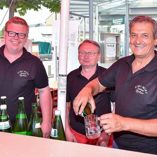 Drei Männer in dunklen Hemden lächeln und stehen hinter einer Bar. Der Mann in der Mitte gießt ein Getränk in ein Glas. Dahinter stehen mehrere Flaschen und ein Pfosten.