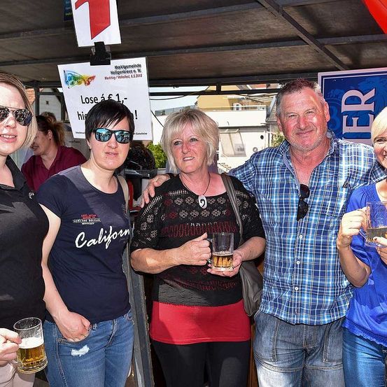 Fünf Menschen stehen bei einer Veranstaltung im Freien zusammen. Eine Frau in der Mitte hält ein Glas Bier. Eine andere Frau auf der linken Seite trägt ein California-Shirt. Hinter ihnen steht ein Schild mit dem Wort 'Lose'.