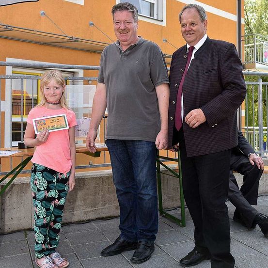 Drei Menschen stehen draußen; ein junges Mädchen hält ein Zertifikat, ein Mann in einem grauen T-Shirt und ein Mann im Anzug. Hinter ihnen sitzt eine weitere Person auf einer Bank.