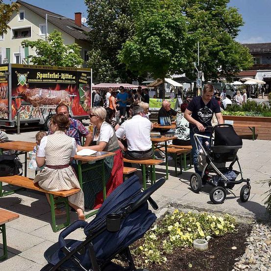 Ein Freiluftmarkt mit Ständen und Menschen, die an Tischen sitzen. Ein Mann schiebt einen Kinderwagen. Es gibt Bäume und Pflanzen in der Umgebung. Ein LKW mit einem Schild für Spanferkel-Ritter.