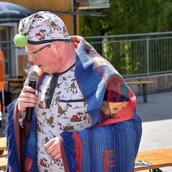 Ein Mann mit Brille und einem bunten Schal hält ein Mikrofon in die Hand und spricht, möglicherweise bei einer Veranstaltung im Freien.