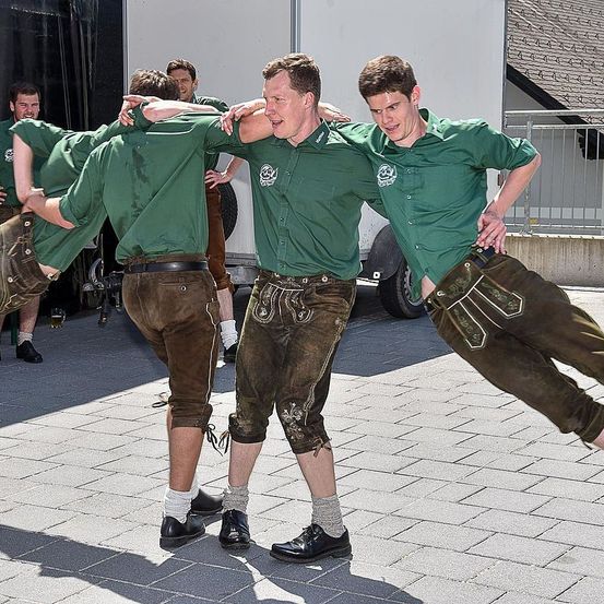Gruppe von Männern, gekleidet in traditionelle bayerische Kleidung, einschließlich grüner Hemden und brauner Lederhosen, führt einen Tanz auf einem Ziegelpflaster im Freien auf.