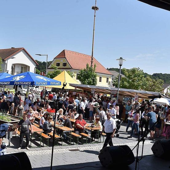 Ein Außenbereich voller Menschen, Tische, Bänke und Sonnendächer. Einige Leute sitzen und essen, andere stehen und gehen. Ein Mann in Weiß geht an einem Lautsprecher auf dem Boden vorbei. Bäume und Gebäude im Hintergrund.