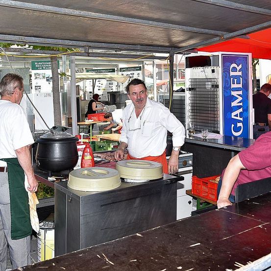 Ein Mann in einer Schürze kocht in einem großen Topf an einem Imbissstand. Ein anderer Mann steht neben ihm, und eine Frau sitzt vor der Theke.