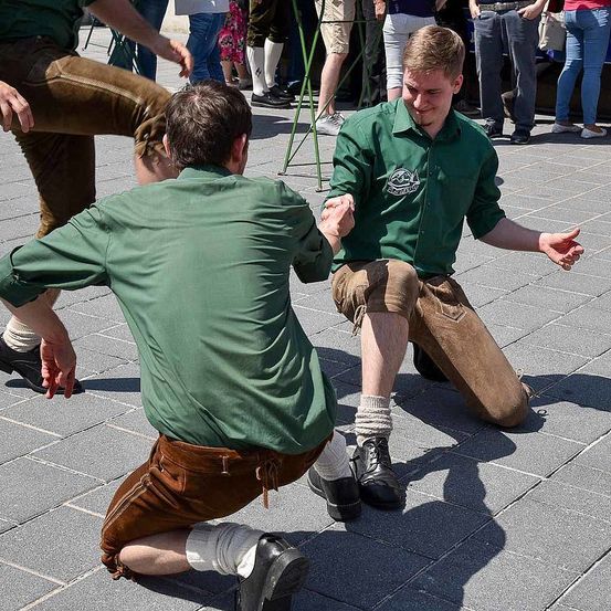 Zwei Männer in grünen Hemden und braunen Hosen knien und halten Händchen in einem Außenbereich. Andere Leute in verschiedenen Outfits stehen und gehen im Hintergrund.