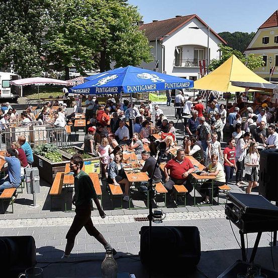 Ein überfüllter Außenbereich mit Tischen und Stühlen, Menschen beim Essen, einer Person, die geht, und großen Schirmen und Zelten. Bäume und Gebäude im Hintergrund.