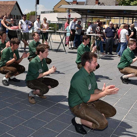 Eine Gruppe von Menschen in grünen Hemden und braunen Hosen kniet und klatscht im Freien. Hinter ihnen stehen und gehen mehrere Personen herum, und es gibt Tische mit Gegenständen darauf.