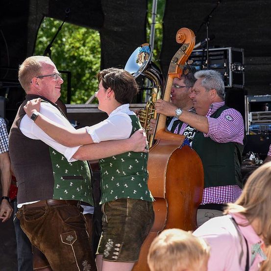 Eine Gruppe von Menschen tanzt und lächelt auf der Bühne. Hinter ihnen hält ein Mann ein großes Cello und ein anderer spielt Tuba. Sie sind alle in traditioneller deutscher Kleidung gekleidet.