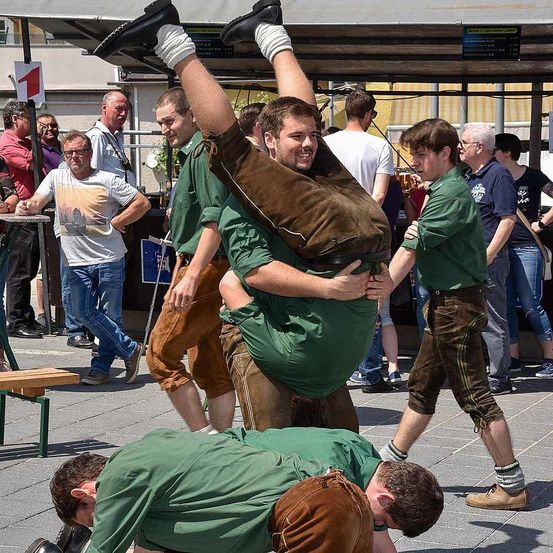 Eine Gruppe von Männern in grünen Shirts und braunen Hosen führt eine traditionelle Hebeaktion auf einem öffentlichen Platz durch. Ein Mann wird von zwei anderen gehoben, während andere zusehen und sich unterhalten.