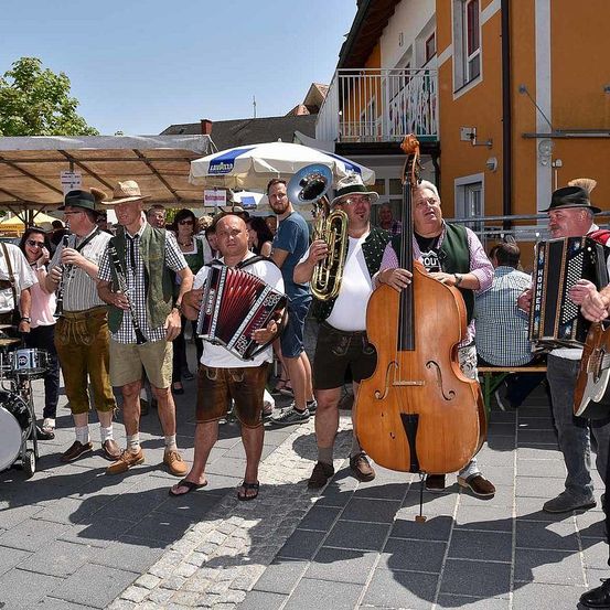 Eine Gruppe von Männern spielt auf der Straße Instrumente. Einige spielen Akkordeons, andere spielen Cello und Schlagzeug. Sie tragen traditionelle bayerische Kleidung.