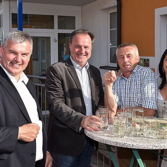 Vier Männer stehen um einen Tisch mit Gläsern Wasser, lächeln und einer zeigt mit dem Finger. Dahinter steht ein Gebäude mit Glasfenstern und einer Tür.