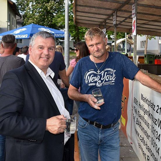 Zwei Männer stehen unter einem Vordach und halten Getränke in den Händen. Einer trägt ein blaues Shirt mit New York, der andere einen dunklen Anzug. Sie lächeln für die Kamera.