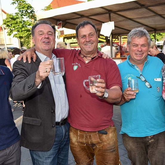 Vier Männer lächeln und halten Gläser mit Bier. Einer trägt ein weinrotes Hemd und braune Hose, ein anderer ein blaues Hemd. Ein Mann in einem Anzug steht mit seinem Arm um den Mann im weinroten Hemd.