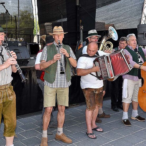 Eine Gruppe älterer Männer spielt verschiedene Musikinstrumente, darunter Klarinette, Akkordeon und Posaune, auf einer Bühne. Sie sind in traditioneller Kleidung gekleidet.