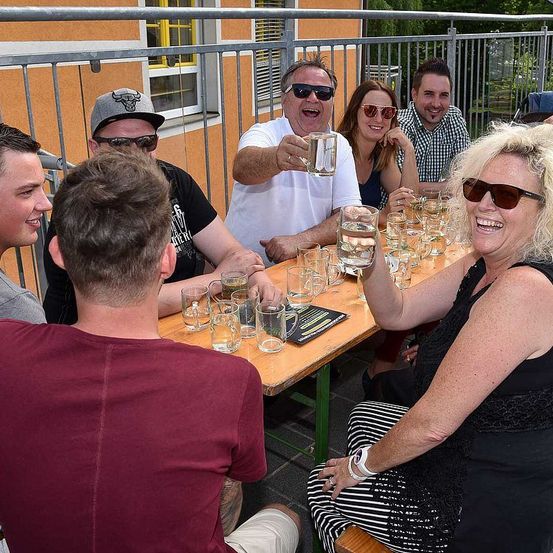 Eine Gruppe von Erwachsenen sitzt um einen Tisch herum und trinkt Gläser Wasser, möglicherweise auf einer Terrasse. Hinter ihnen befindet sich ein Metallgeländer. Die Frau rechts lächelt.