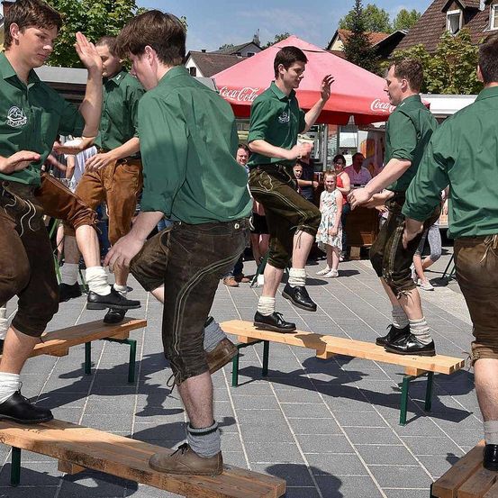 Eine Gruppe junger Männer in grünen Hemden und braunen Hosen führt einen Tanz auf hölzernen Bänken im Freien auf. Ein Coca-Cola-Schirm ist im Hintergrund zu sehen.