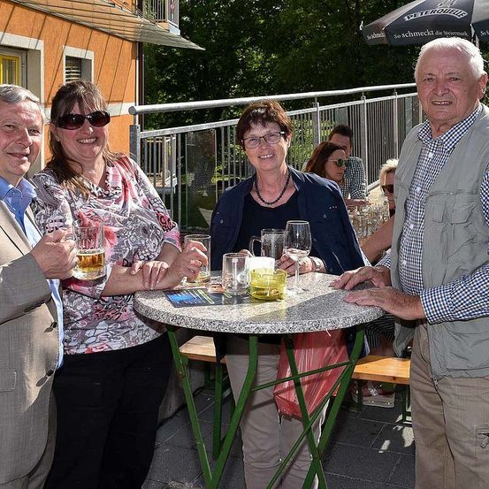 Vier Personen stehen um einen Tisch in einem Outdoor-Restaurant und halten Gläser mit Bier. Sie lächeln und posieren für ein Foto. Hinter ihnen befindet sich ein Metallgeländer und in der Ferne sitzen und stehen weitere Personen.