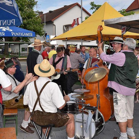 Eine Gruppe von Männern in traditioneller deutscher Kleidung spielt Musikinstrumente auf einer Straße. Sie sind von einer Menge Menschen und gelben Zelten umgeben. Ein Flagge ist im Hintergrund zu sehen.