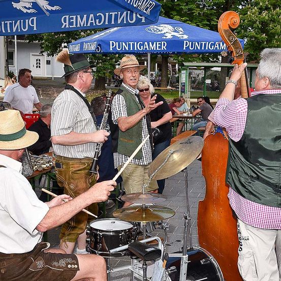 Eine Band spielt im Freien auf einem gepflasterten Bereich mit blauen Sonnenschirmen. Männer spielen Schlagzeug, Cello und Klarinette. Menschen sitzen an Tischen unter den Sonnenschirmen.