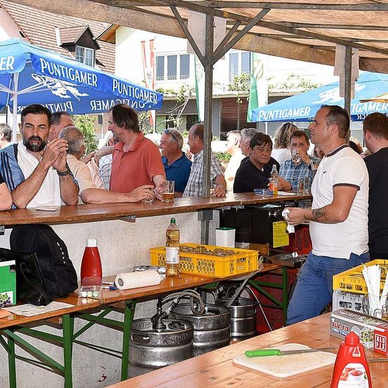 Eine Gruppe von Männern sitzt unter einem Vordach an einer Bar, einige trinken. Ein Mann in Weiß reinigt die Theke. Flaschen, Gläser und Fässer sind in der Nähe.