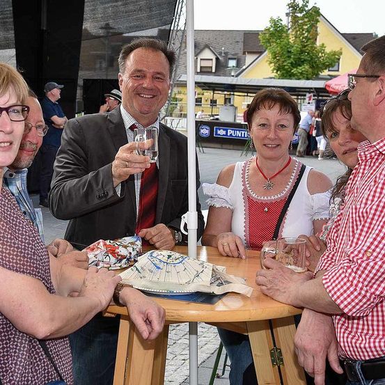 Mehrere Menschen versammeln sich um einen Tisch, lächeln und halten Gläser in den Händen. Ein Mann im Anzug hebt ein Glas, während eine Frau in einem weißen Oberteil mit rotem Rand lächelt. Hinter ihnen steht ein großer Schirm, mit einem Schild, auf dem 'PUNT' steht. Im Hintergrund sind Gebäude und Bäume zu sehen.