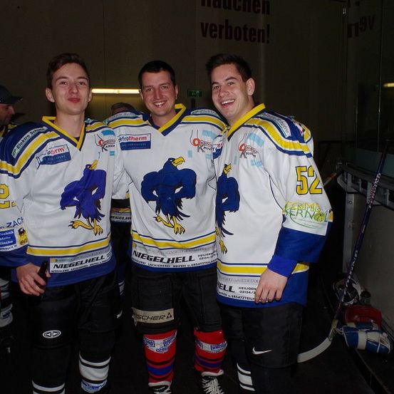 Drei Eishockeyspieler stehen in einem Umkleideraum, lächeln und posieren für ein Foto. Sie tragen gleiche Trikots mit demselben Logo und der Nummer 52 auf dem Rücken. Sie sind umgeben von Eishockeyschlägern, Handschuhen und einem Schild, auf dem steht 'Rauchen verboten.'