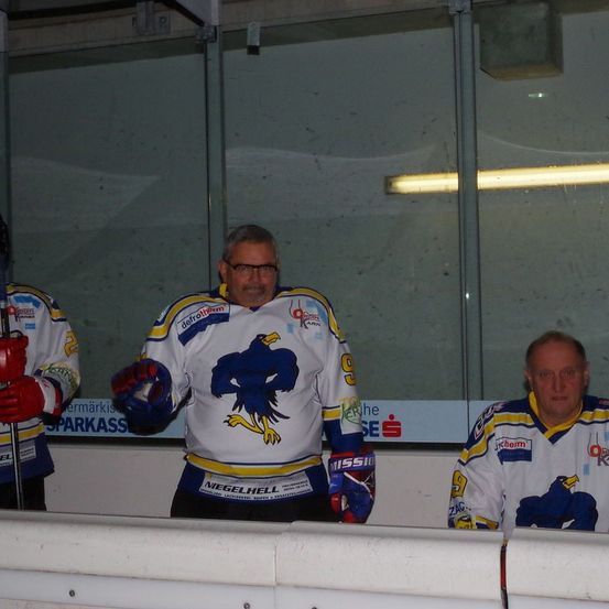 Drei Männer sitzen auf einer Bank in einer überdachten Eishockeyhalle. Der Mann in der Mitte trägt ein Trikot mit blauem Adlerdesign. Die beiden Männer an den Seiten tragen rote Handschuhe.