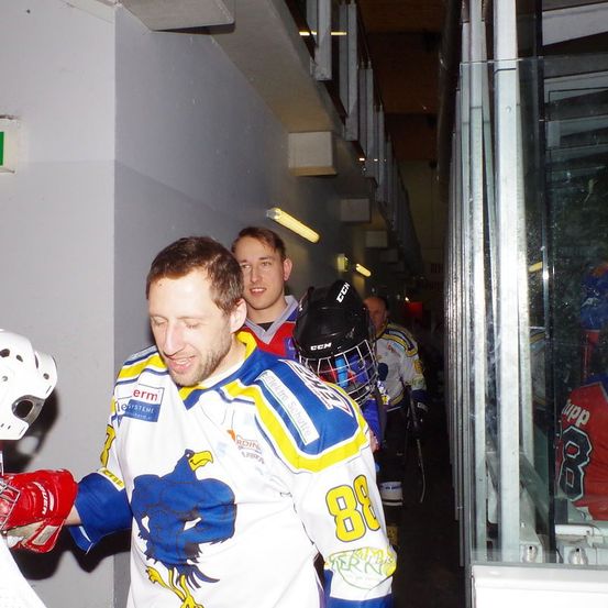 Ein Eishockey-Spieler mit einem Trikot mit der Nummer 88 lächelt, während er die Umkleidekabine verlässt. Ein Spieler mit Helm steht hinter ihm.