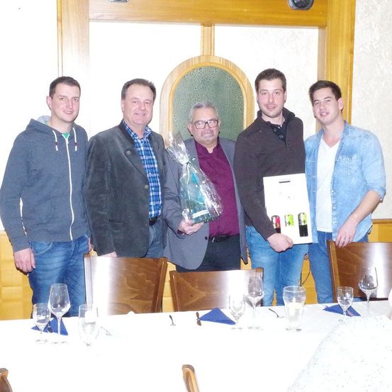 Fünf Männer stehen in einem Restaurant an einem Tisch und lächeln. Ein Mann hält ein eingewickeltes Geschenk, ein anderer ein gerahmtes Bild und die anderen halten Weingläser. Hinter ihnen befindet sich ein Fenster und eine weiße Wand.