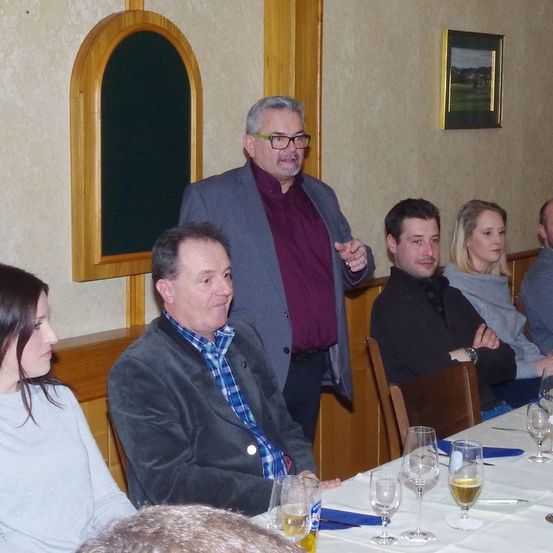 Eine Gruppe von Menschen sitzt in einem Restaurant um einen Tisch herum, ein Mann steht und gestikuliert, während er spricht, ein anderer sieht zu und hat die Arme verschränkt.
