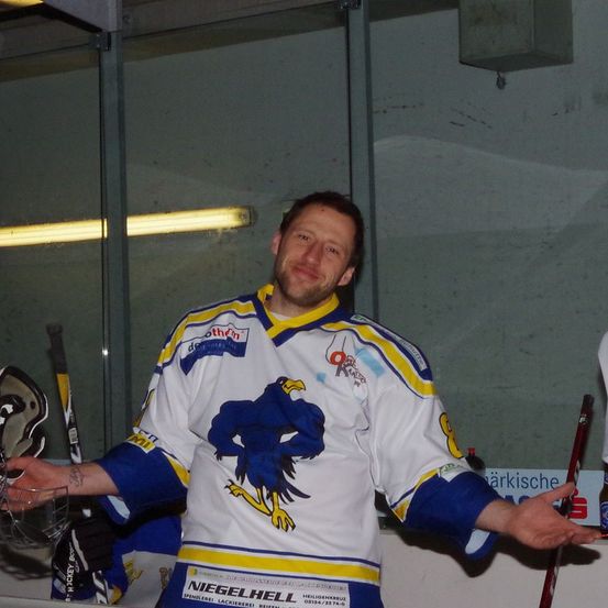 Ein Eishockeyspieler steht lächelnd in einer Arena und trägt ein weißes Trikot mit einem blauen Adler und gelben Besatz. Er hält einen Eishockeyschläger in der rechten Hand und posiert mit ausgestrecktem linken Arm.
