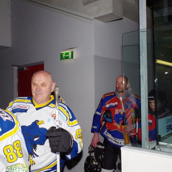 Drei Eishockeyspieler stehen im Flur einer Arena. Einer hält einen Schläger. Ein Schild über der Tür steht 'Ausgang'. Eine Glaswand reflektiert die Spieler.