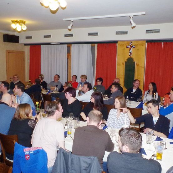 Eine Gruppe von Menschen sitzt in einem Speisesaal an Tischen, einige von ihnen trinken. Auf den Tischen stehen Weingläser, Teller und Servietten. Der Raum hat weiße Vorhänge, eine Holztür und eine Holzwand mit einem Kreuzsymbol.