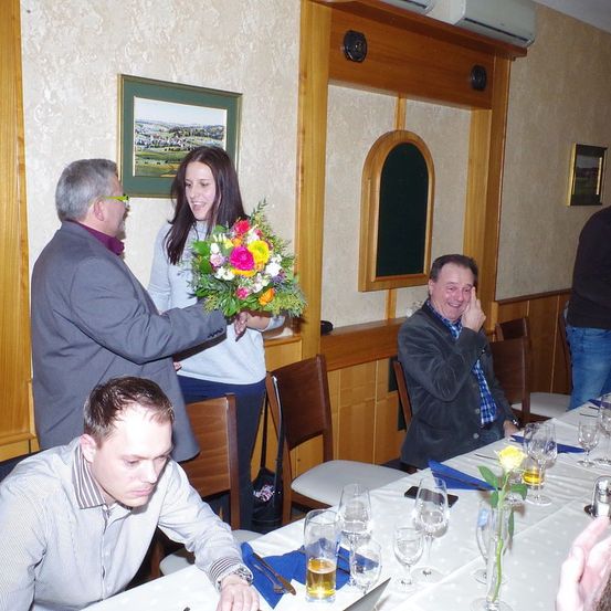 Ein Mann überreicht einer lächelnden Frau, die neben ihm steht, einen Blumenstrauß. Ein sitzender Mann lacht, während andere um einen Tisch mit Getränken herumsitzen.