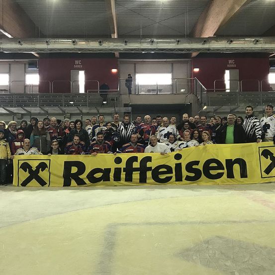 Eine große Gruppe von Menschen, einschließlich Hockeyspielern, steht hinter einem gelben Banner mit dem Wort Raiffeisen und posiert für ein Foto in einer überdachten Arena.