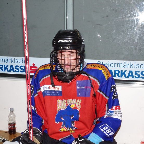 Ein Eishockeyspieler sitzt auf der Bank, trägt einen Helm und hält einen Schläger. Er ist in einem blauen, roten und weißen Trikot mit einem Adlerlogo und dem Wort Blue Eagles auf der Vorderseite gekleidet.