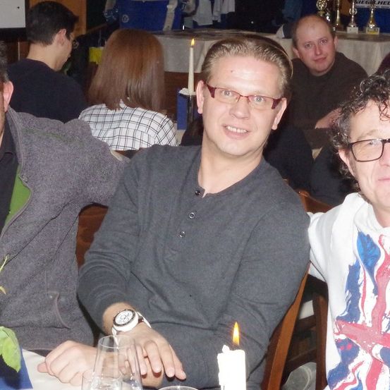 Mehrere Personen sitzen um einen Tisch herum, ein Mann trägt eine Brille und eine Uhr, und ein anderer Mann trägt ein T-Shirt mit Union Jack-Design. Sie lächeln und scheinen für ein Foto zu posieren. Es gibt Kerzen auf dem Tisch. Hinter ihnen sitzen noch mehr Leute, und auf einem Tisch stehen Trophäen.