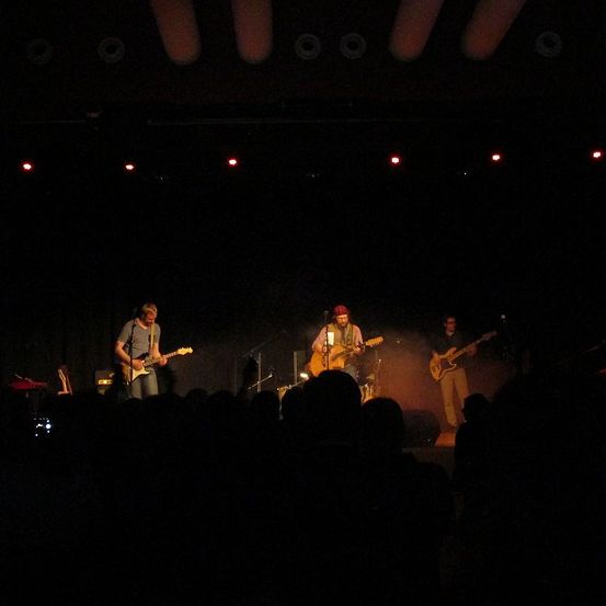 Eine Band tritt bei Nacht auf der Bühne auf, beleuchtet von roten Lichtern. Drei Musiker spielen Gitarren, einer trägt einen roten Hut. Das Publikum sitzt im Dunkeln und konzentriert sich auf die Aufführung.