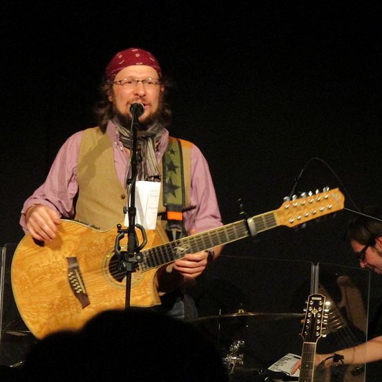 Ein Mann mit rotem Bandana und Brille spielt Gitarre auf der Bühne vor einem Publikum.