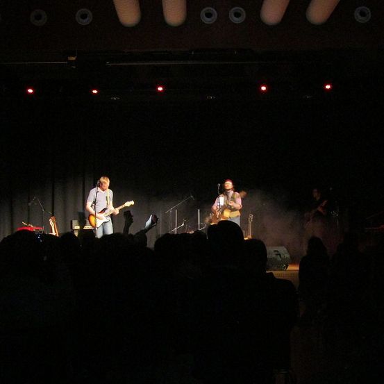 Eine Band spielt auf der Bühne unter roten Lichtern, wobei zwei Gitarristen im Rampenlicht stehen, während ein Publikum unten zuschaut.